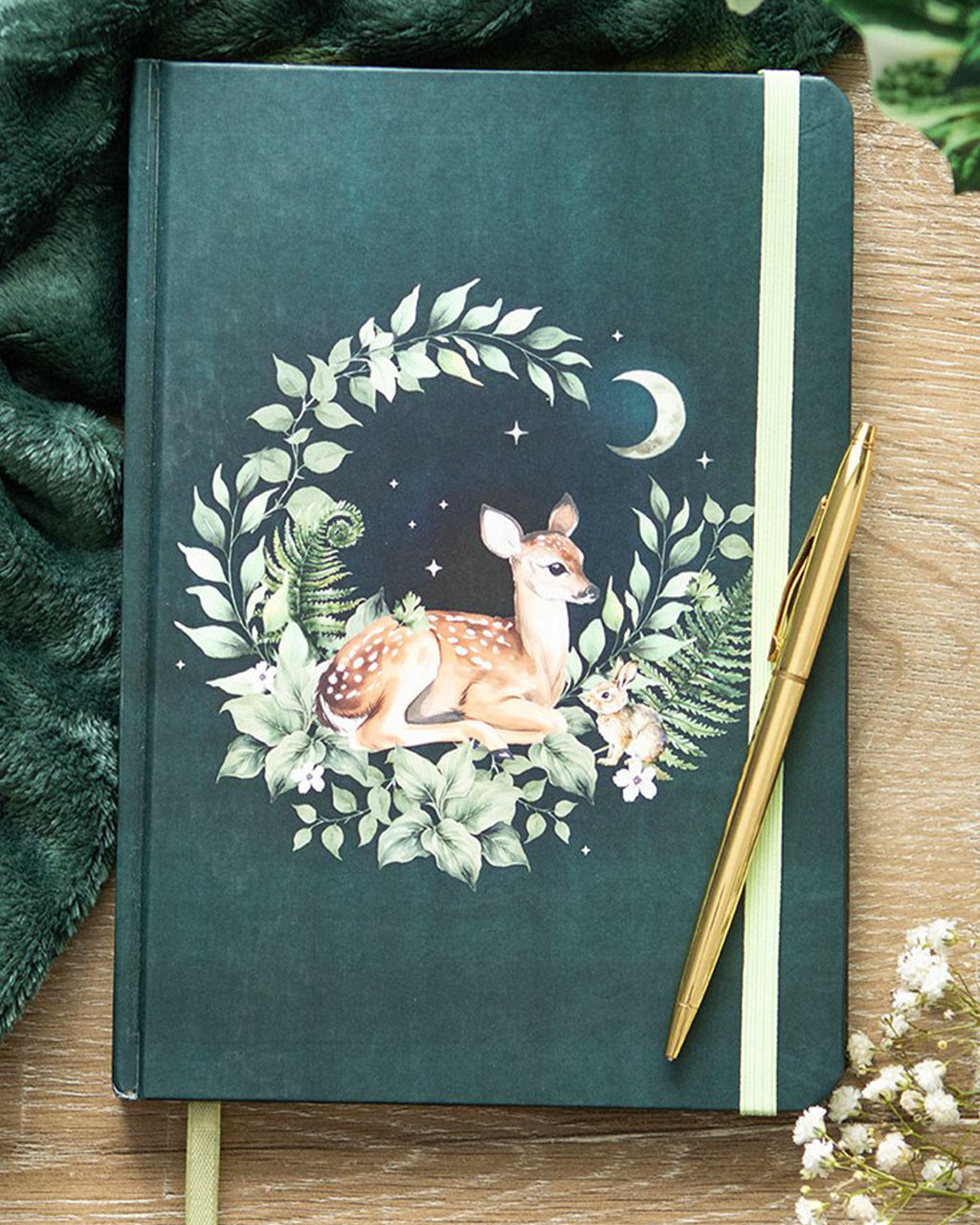 A5 notebook with a forest green cover showcasing a delicate fawn surrounded by foliage, flowers, and a crescent moon. Finished with gold-edged pages and a green elastic closure.