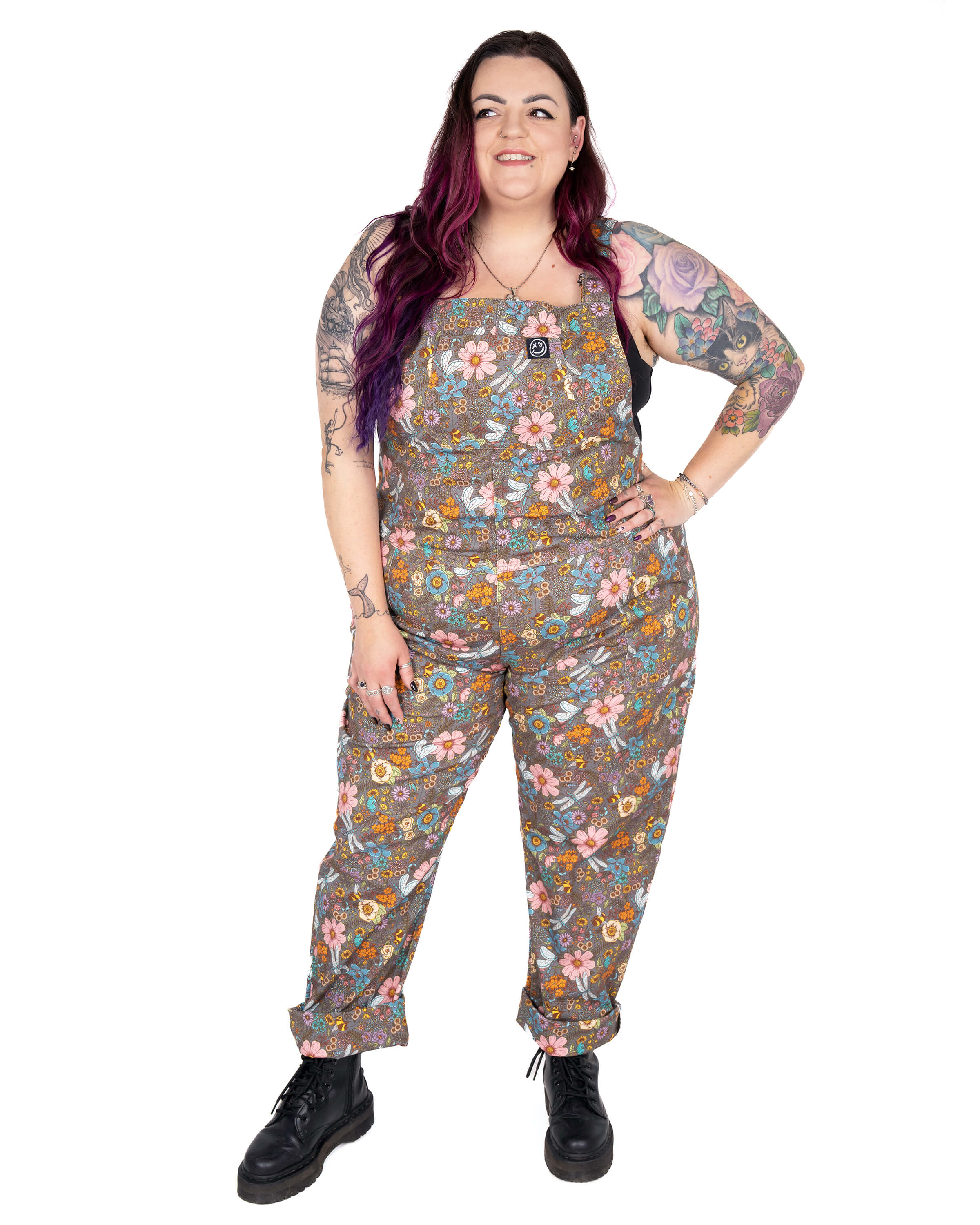 Luisa wears Rebel Romance Cottagecore Stretch Twill Dungarees, styled with rolled-up cuffs and chunky black boots. The floral design includes pink, blue and yellow flowers with dragonflies on a brown base. Luisa is a UK 22/24, 5'6