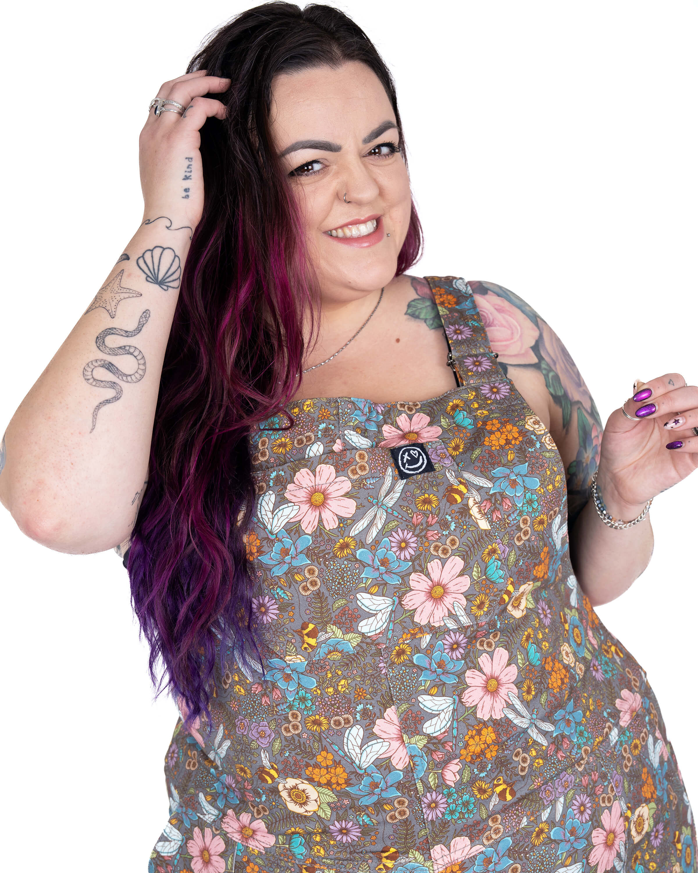Luisa wears Rebel Romance Cottagecore Stretch Twill Dungarees featuring a whimsical floral and dragonfly print in pastel tones. She has long purple ombré hair and visible tattoos, smiling confidently with one hand in her hair. Luisa is a UK size 22/24 wearing a 3XL. Rebel Romance is a small UK brand celebrating alternative slow fashion and bold style for millennials.