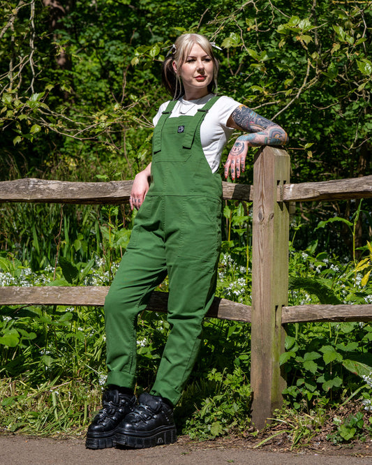 Model wearing Forest Green Stretch Twill Dungarees leaning against a wooden fence in the sunshine, surrounded by greenery.