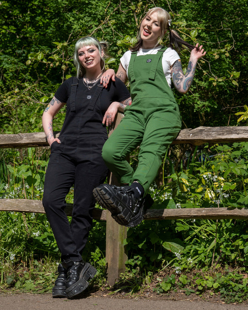 Models wearing Forest Green and Jet Black Stretch Twill Dungarees by Rebel Romance, seated together on a fence in dappled sunlight.