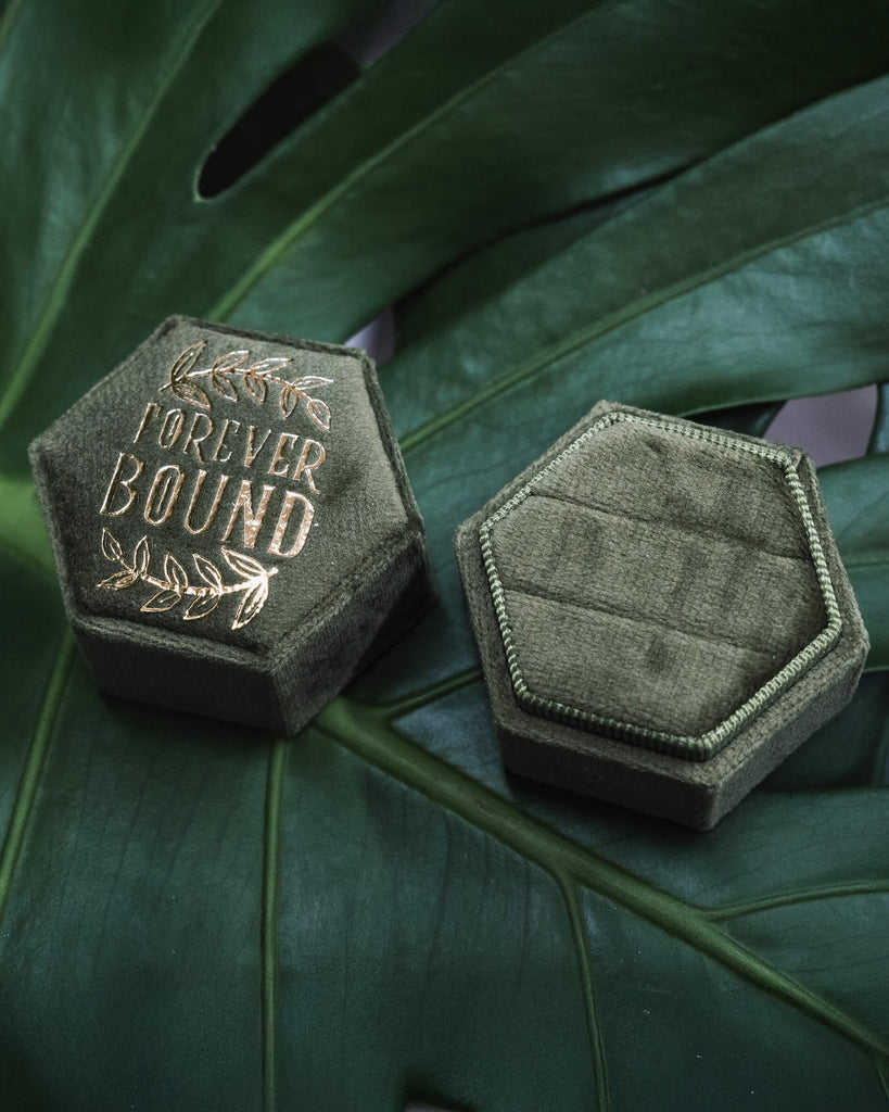 A dark green hexagonal velvet ring box embossed with the words “Forever Bound” in gold foil surrounded by delicate leaf illustrations. The box rests on a lush green monstera leaf.
