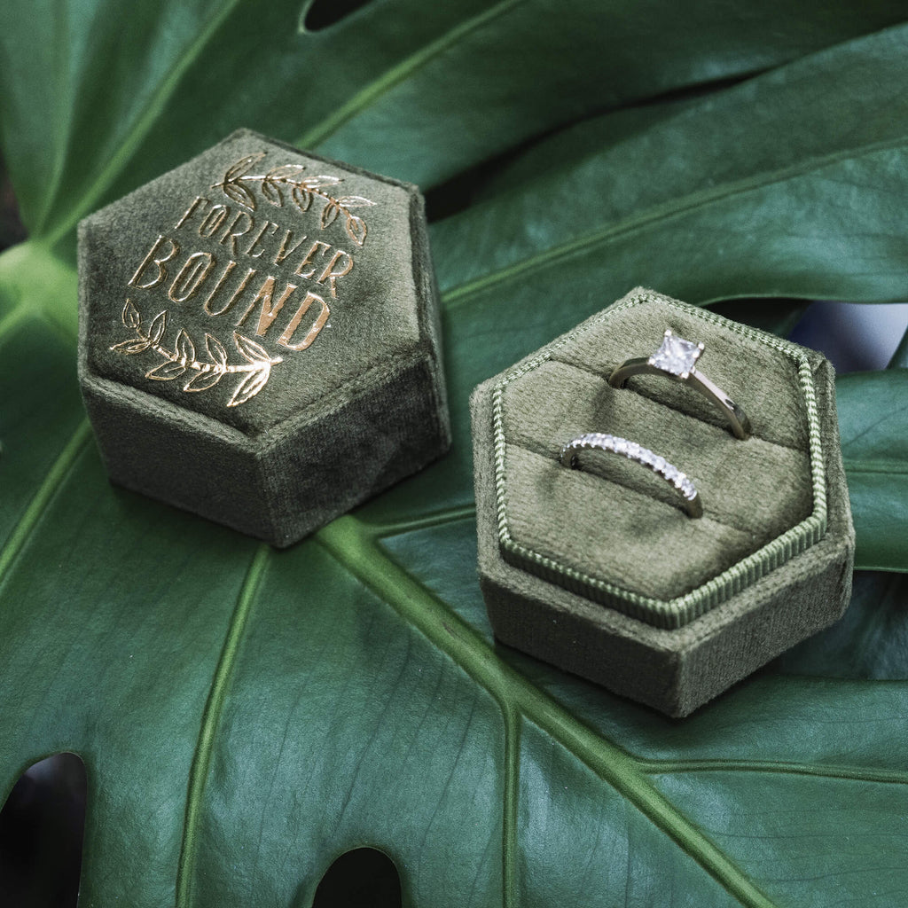  Two halves of the “Forever Bound” hexagonal velvet ring box displayed open on a green monstera leaf, showcasing two rings inside—a solitaire engagement ring and a matching diamond band. The box is deep green with gold foil text and leaf accents.