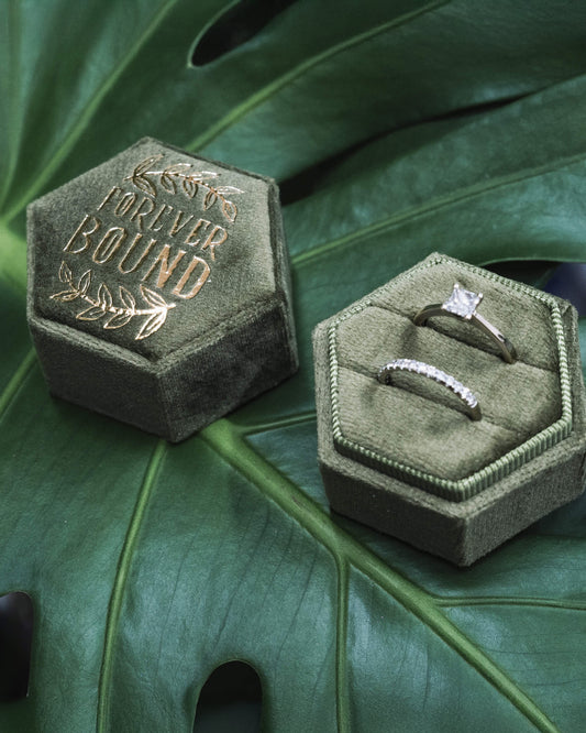  Two halves of the “Forever Bound” hexagonal velvet ring box displayed open on a green monstera leaf, showcasing two rings inside—a solitaire engagement ring and a matching diamond band. The box is deep green with gold foil text and leaf accents.