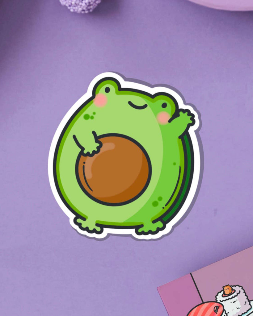 A cute vinyl sticker of a green cartoon frog with rosy cheeks, shaped like an avocado with a large brown stone in the centre. The sticker is shown on a purple background. Ideal for decorating laptops, water bottles, notebooks and more.