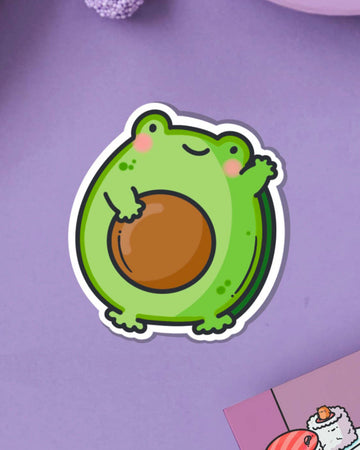 A cute vinyl sticker of a green cartoon frog with rosy cheeks, shaped like an avocado with a large brown stone in the centre. The sticker is shown on a purple background. Ideal for decorating laptops, water bottles, notebooks and more.
