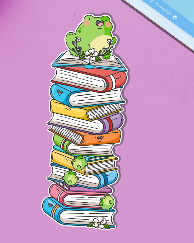A cute rectangular bookmark showing a cheerful green frog sitting on top of a tall, colourful stack of cartoon books with smiling faces. Printed on glossy card with flowers and tiny frogs tucked between the books. A playful accessory for readers and stationery lovers.