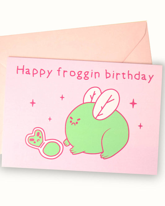 A pastel pink greeting card featuring an illustration by Nimbob of a round, grumpy green frog with soft pink fairy wings. The frog has a pouty expression and is staring at a small frog-shaped compact mirror lying on the ground. Sparkles and whimsical details surround the illustration. Above, the text reads ‘Happy Froggin’ Birthday’ in a playful, handwritten font. A fun and cheeky birthday card, perfect for frog lovers and those with a sassy sense of humour