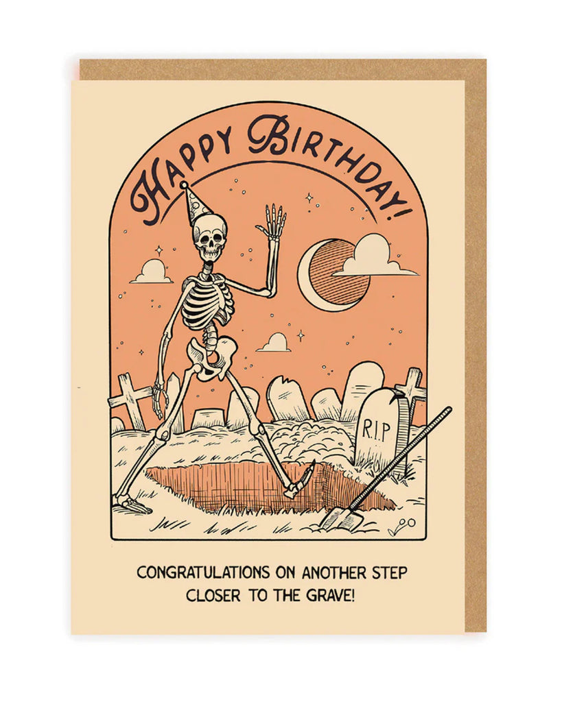 A front-facing shot of the skeleton birthday card, standing upright against a black backdrop. The fun and eerie design features a skeleton in a playful pose, surrounded by a graveyard scene, with the cheeky birthday message in bold letters. The card is paired with a brown kraft envelope for a rustic finish.
