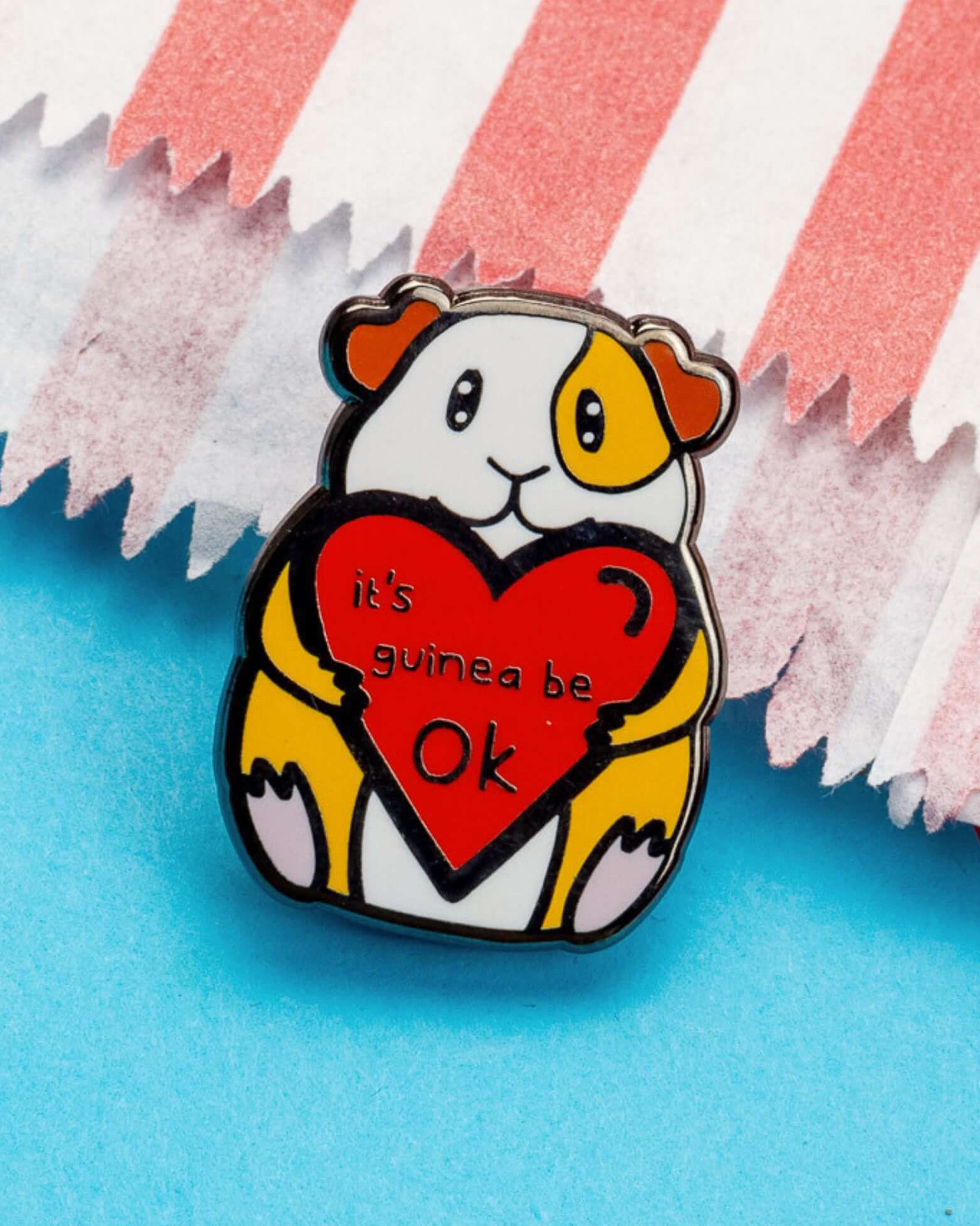 Close-up image of the 'It's Guinea Be Ok' enamel pin by Innabox, sold by Rebel Romance. The pin shows an adorable guinea pig holding a heart with the phrase “It’s guinea be ok” written on it. Background includes red, white, and blue paper textures. A perfect gift for guinea pig lovers and those in need of a little emotional boost.