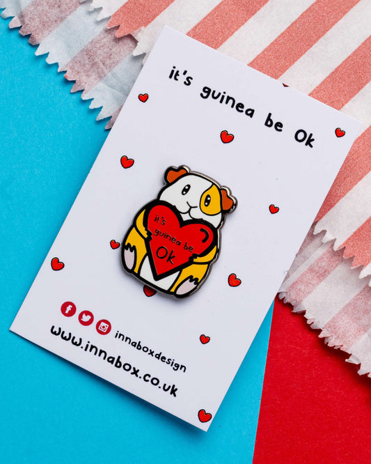 Product photo of the 'It's Guinea Be Ok' enamel pin by Innabox, available at Rebel Romance. The pin features a cute guinea pig holding a red heart with the punny message "It's guinea be ok", displayed on backing card with tiny red hearts. Surrounded by blue and red paper stripes. Designed to raise mental health awareness with a cute and comforting twist.