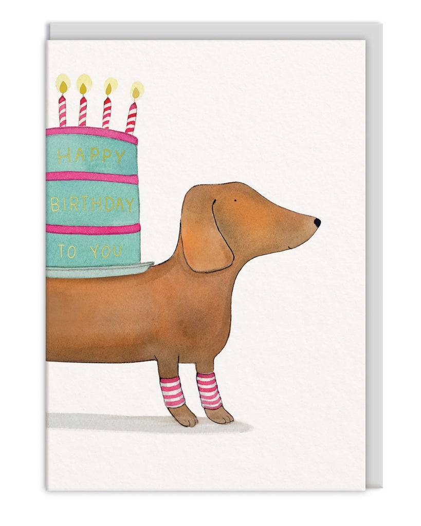 Cute birthday greeting card featuring a dachshund wearing pink striped socks carrying a pastel blue and pink birthday cake with candles and gold foil lettering reading Happy Birthday To You Comes with a grey envelope Perfect for dog lovers and birthdays
