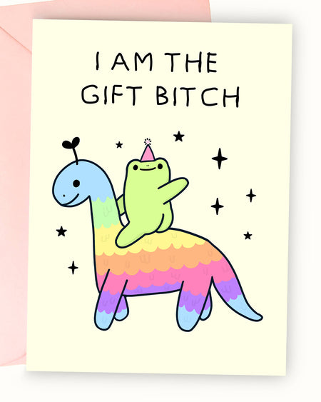 A pastel yellow greeting card featuring an illustration by Nimbob of a cute green frog sitting on a rainbow-coloured dinosaur. The frog wears a tiny pink party hat and raises one arm triumphantly. Above, the bold black text reads ‘I AM THE GIFT BITCH.’ The dinosaur has a friendly face with a tiny heart-shaped antenna, and the background includes scattered sparkles and stars. A fun and sassy birthday card perfect for celebrating in style!