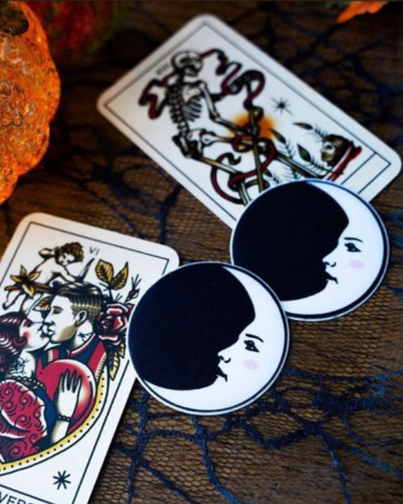 Two Moon: Black and White Man in the Moon Nipple Pasties by Pastease shown next to two tarot cards and glass pumpkins on a black net table cover.