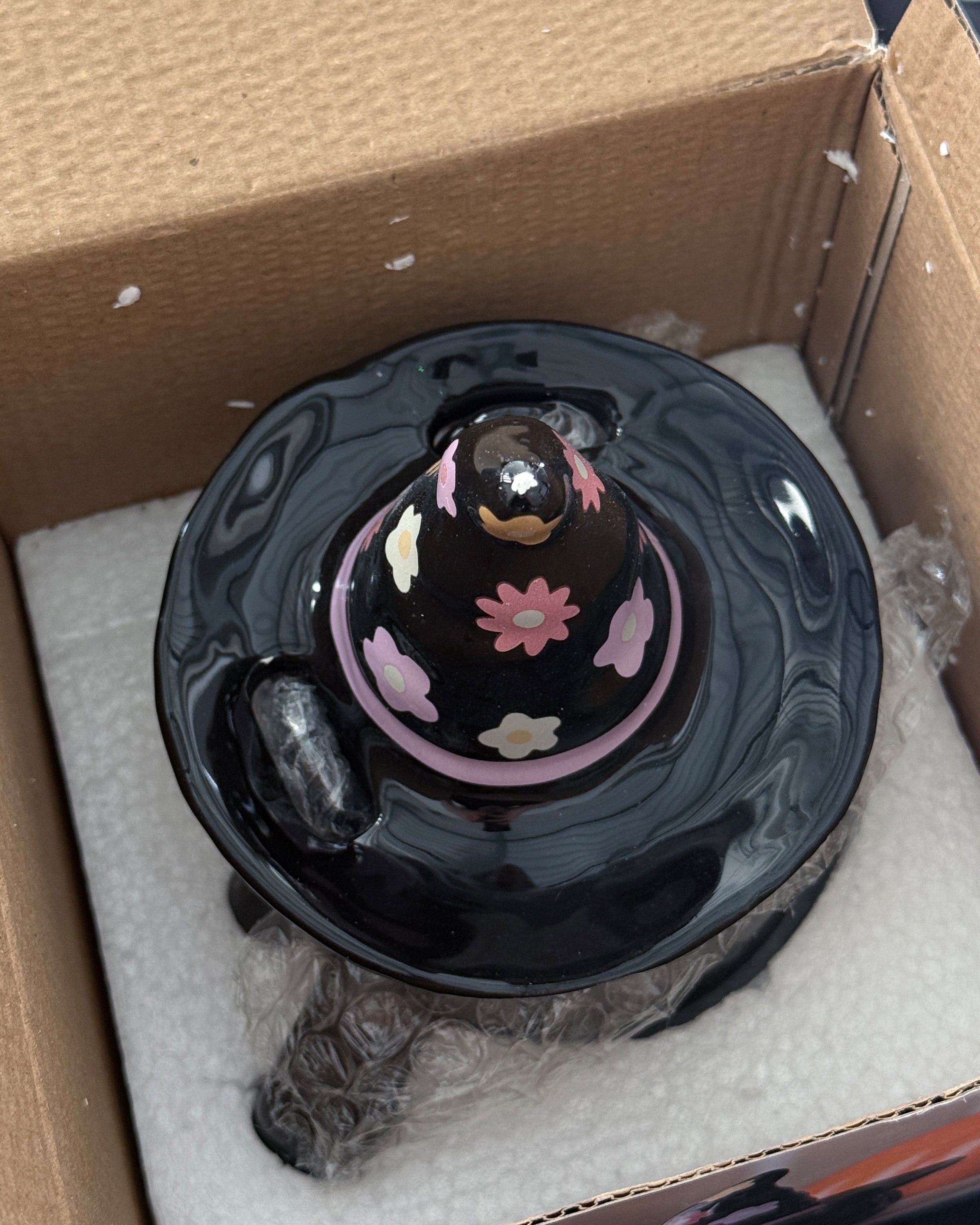 Rebel Romance Groovy Witch Cat Lidded Mug inside its protective packaging with a tiny chip off the top of the hat. The black ceramic mug has a removable witch hat lid with pink, yellow and white flower designs. A whimsical, gothic-inspired homeware piece perfect for alternative style lovers, Halloween fans and millennials who enjoy quirky, magical mugs. A unique gift idea combining fun, spooky charm with everyday functionality.