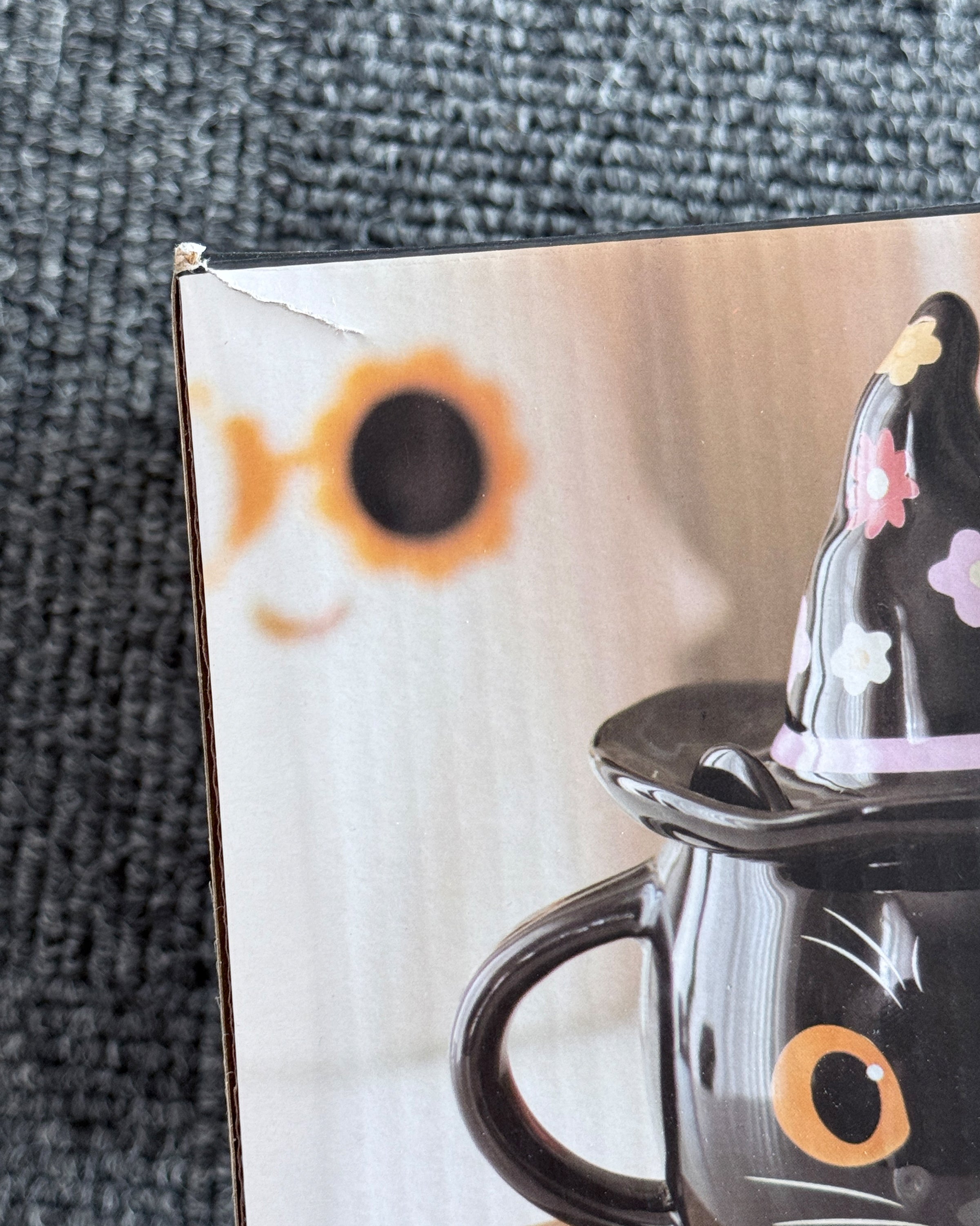 Close-up of the Rebel Romance Groovy Witch Cat Lidded Mug packaging that has a small tear, featuring a black ceramic cat mug with big orange eyes and a removable witch hat lid decorated with colourful flowers. The alternative homeware gift box has a small tear on the corner. Perfect gothic and quirky kitchenware for millennials who love witchy, Halloween-inspired mugs and unique ceramic drinkware.