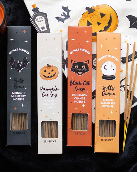 A Halloween-themed incense stick gift set featuring four boxes labelled Pumpkin Carving, Spells Divine, Bat’s Brew, and Black Cat Curse. Each box contains 15 sticks with seasonal scents like spiced pumpkin, mystic mandarin, midnight mulberry, and cinnamon orange. Surrounded by Halloween decor including pumpkins, bats, and a candle on a cosy black fabric background.