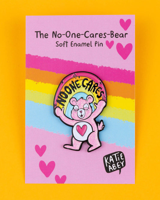 The "No-One Cares Bear" pin by Katie Abey features a pink glitter bear with a heart on its belly and a rainbow that reads "No-One Cares." Displayed on a bright pink backing card with rainbow stripes and hearts, the pin is full of humour and attitude. This playful soft enamel pin makes a bold statement about not caring, encouraging you to embrace your quirky side. Katie Abey’s unique style mixes fun and weirdness, making it a great accessory for jackets, bags, or gifts.