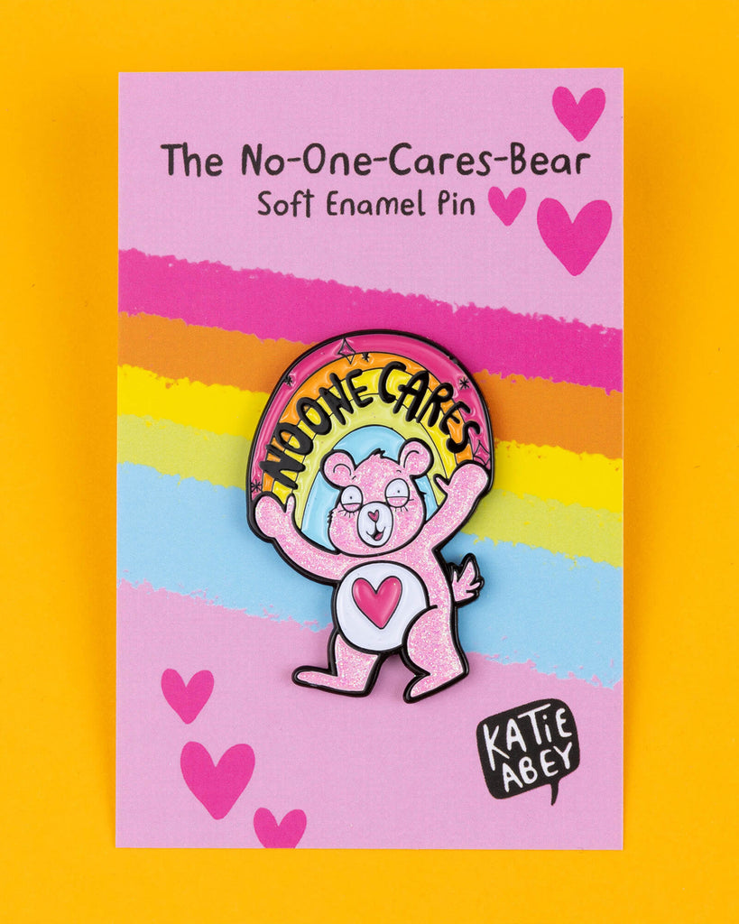 The "No-One Cares Bear" pin by Katie Abey features a pink glitter bear with a heart on its belly and a rainbow that reads "No-One Cares." Displayed on a bright pink backing card with rainbow stripes and hearts, the pin is full of humour and attitude. This playful soft enamel pin makes a bold statement about not caring, encouraging you to embrace your quirky side. Katie Abey’s unique style mixes fun and weirdness, making it a great accessory for jackets, bags, or gifts.