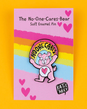 The "No-One Cares Bear" pin by Katie Abey features a pink glitter bear with a heart on its belly and a rainbow that reads "No-One Cares." Displayed on a bright pink backing card with rainbow stripes and hearts, the pin is full of humour and attitude. This playful soft enamel pin makes a bold statement about not caring, encouraging you to embrace your quirky side. Katie Abey’s unique style mixes fun and weirdness, making it a great accessory for jackets, bags, or gifts.