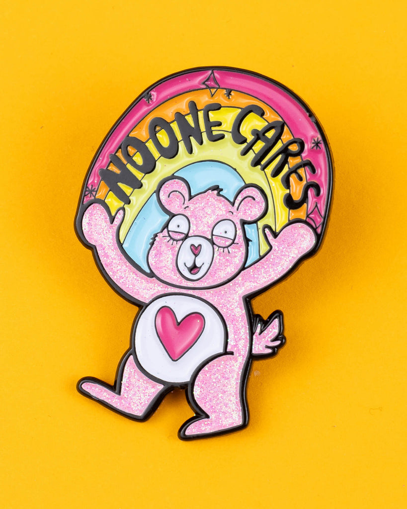 Katie Abey’s "No-One Cares Bear" pin features a pink glitter bear with tired eyes and a heart on its belly, holding a rainbow banner that reads "No-One Cares." The bear is set against a bright yellow background, highlighting its quirky design. This soft enamel pin represents Katie Abey’s signature weird, sassy style. Perfect for pin collectors or as a cheeky gift, it adds a playful and humorous touch to any accessory. Katie Abey's designs encourage embracing individuality and weirdness.