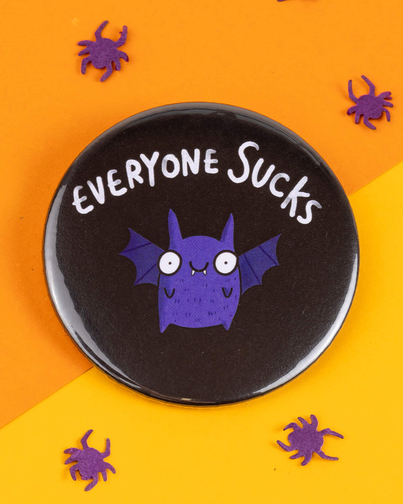 Round 58mm pin-back badge featuring a purple cartoon bat with a cheeky expression and the text "Everyone Sucks" above it. Set against an orange and yellow backdrop with purple spider confetti. Designed by Katie Abey for Rebel Romance, a UK alternative fashion brand.