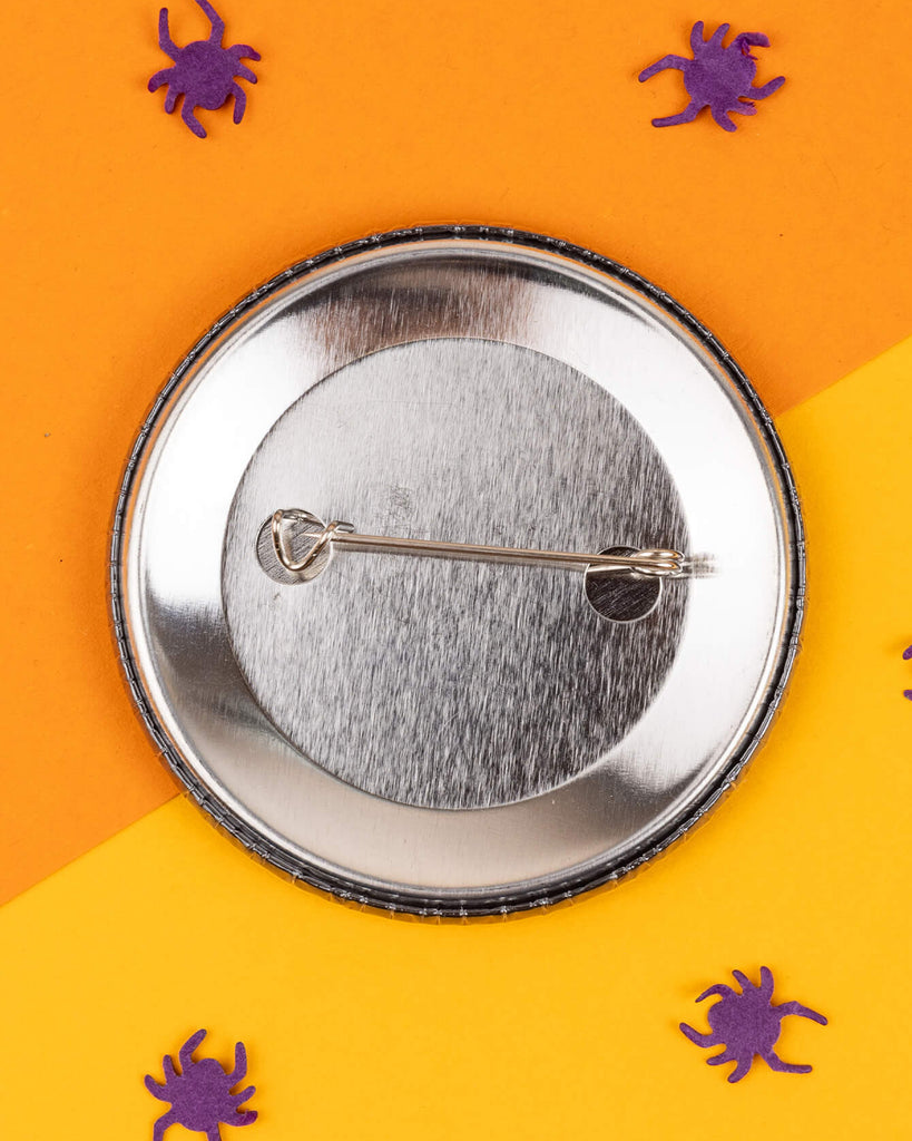 Close-up of the back of a round 58mm pin-back badge with a secure metal clasp, placed on a split orange and yellow background with scattered purple paper spiders. From Rebel Romance, a UK small business celebrating alternative style and aesthetics.
