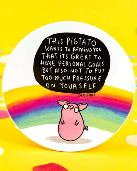 A round vinyl sticker by Katie Abey featuring a pink pig-potato hybrid character called the Pigtato. Text above reads: “This Pigtato wants to remind you that it's great to have personal goals but also not to put too much pressure on yourself.” The background features a soft rainbow on a white base. Durable, motivational, and perfect for water bottles, notebooks, or laptops.