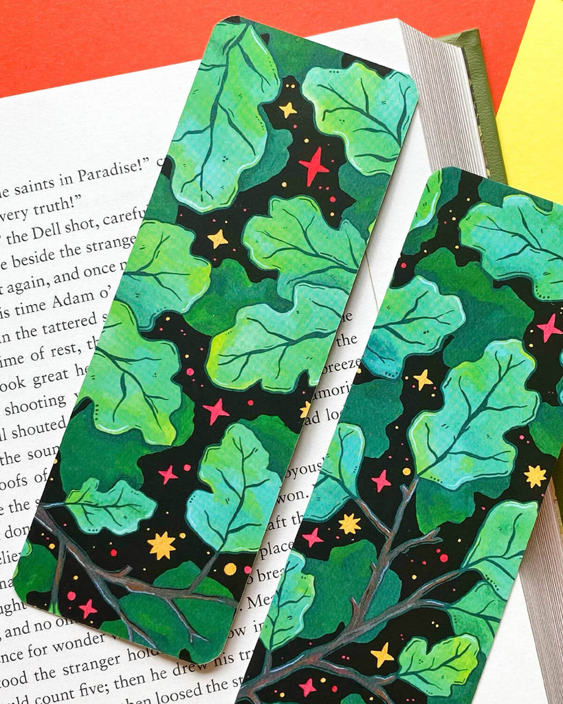  Two bookmarks with a dark background, illustrated with vibrant green leaves, bright stars, and tiny pops of colour in red and yellow. Resting on an open book with warm-toned paper.