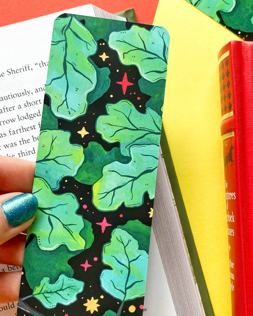 A hand with metallic blue nail polish holding the Deep Dark Woods Bookmark, showing the detailed gouache artwork of green leaves and scattered red and yellow stars on a dark, dreamy background. A book spine and yellow page peek from behind.