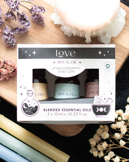 Boxed set of 3 Love Ritual Blended Essential Oils (Thyme, Rose Geranium, Ginger) with celestial packaging on a wooden tray with flowers and candles.