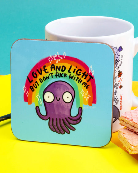 The same rainbow octopus coaster is propped up against a white illustrated mug. The coaster shows a purple cartoon octopus beneath a rainbow with the caption “Love and Light But Don’t Fuck With Me.” It's set on a vibrant yellow surface with a pastel blue backdrop and pink wafers nearby.