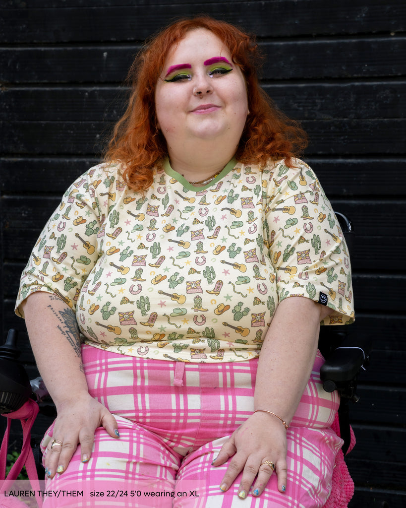 Lauren, a non binary person with long curly ginger hair with colourful bright makeup wearing the Rebel Romance cowboy oversized boxy crop tee. The print is a pastel yellow base with cowboy boots, wanted posters, snakes, cacti and guitars illustrated by Yasmin Cousins with a sage green contrasting neck trim. They've paired them with pink checkerboard trousers and they are smiling sat in their mobility aid with a black door behind. 