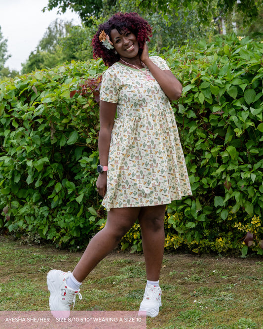 Ayesha, a red and black curly haired person with decorative matching bows, platform white trainers and a pastel A necklace. She is wearing the cowboy smock dress, a pastel butter yellow with doodled cowboy themed illustrations by yasmin cousins and a contrasting neck trim. She is stood outside on grass smiling leaning on one leg and a large bush plant behind her. 