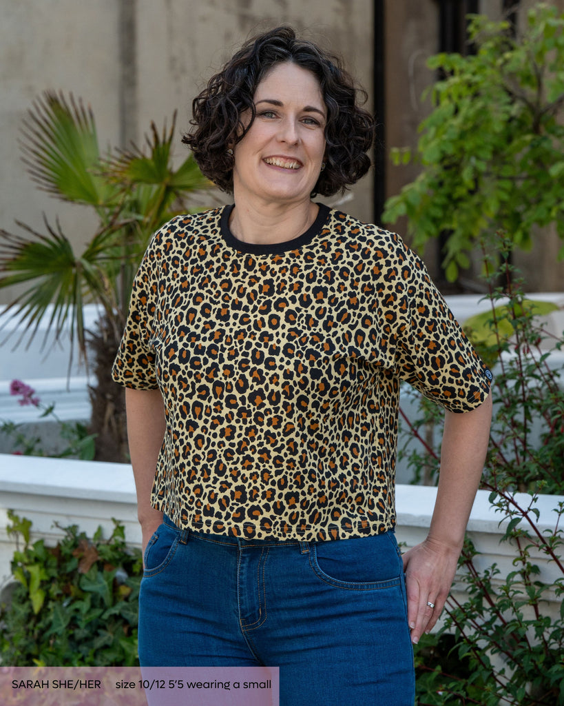 Sarah, a woman with bobbed curly black hair and smokey eye makeup wearing a natural leopard print crop top with a contrasting black neck trim. She's smiling facing forward with her hands on her hips, she's paired it with blue denim jeans stood outside in front of a garden. 