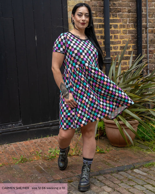 Carmen, a goth model with long black hair posing outside swishing the checkerboard dress she's wearing with silver platform boots. The dress is a black and colourful checkered print with a contrasting black neckline. 