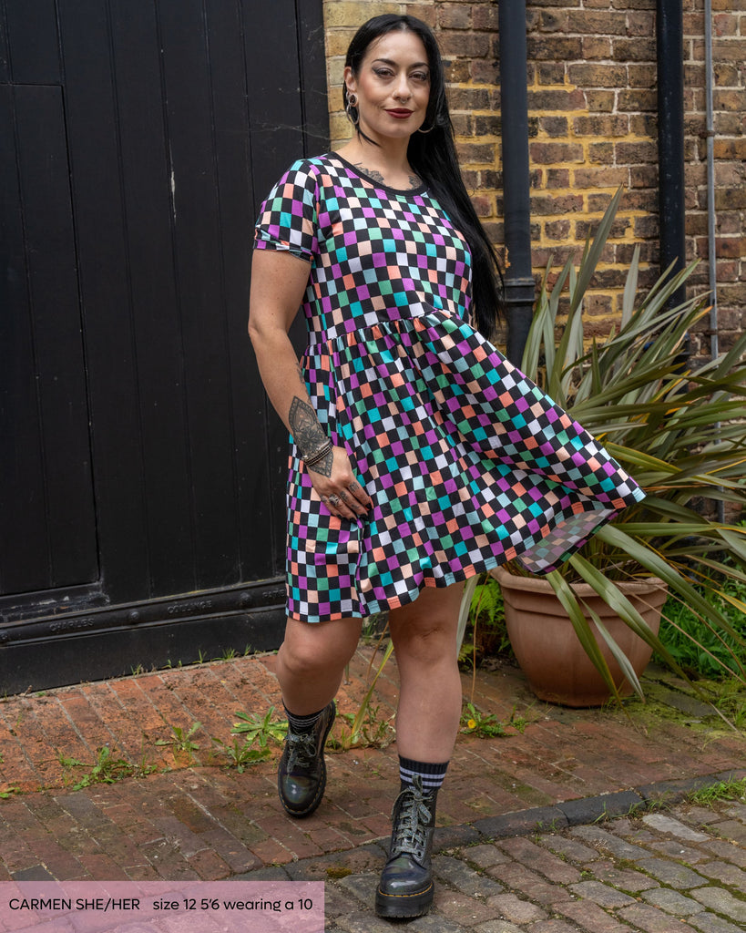 Carmen, a goth model with long black hair posing outside swishing the checkerboard dress she's wearing with silver platform boots. The dress is a black and colourful checkered print with a contrasting black neckline. 