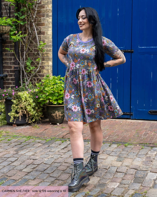 Carmen, a gothic model with long black hair wearing the rebel romance cottagecore smock dress with holographic silver boots. She is taking a step forward with her hands on her hips smiling looking down to the left. The dress is a denim blue base with contrasting purple neck trim, the print features various colourful florals, bees, dragonflies and butterflies. 