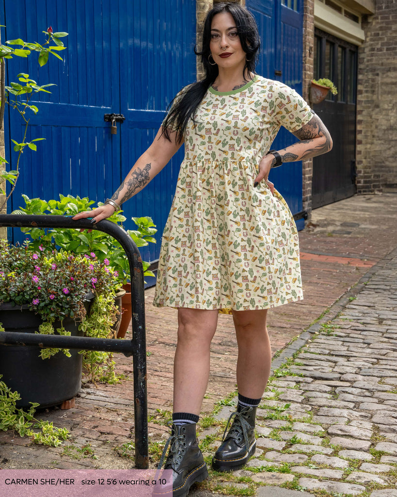 Carmen, a gothic tattooed model with long black hair is wearing the rebel romance x yasmin cousins cowboy smock dress with grey boots. She is facing forward smiling leaning on a black railing with one hand on her hip. The dress print is a pastel soft yellow base with cowboy illustrations all over such as sheriff stars, guitars, cacti, snakes and more.