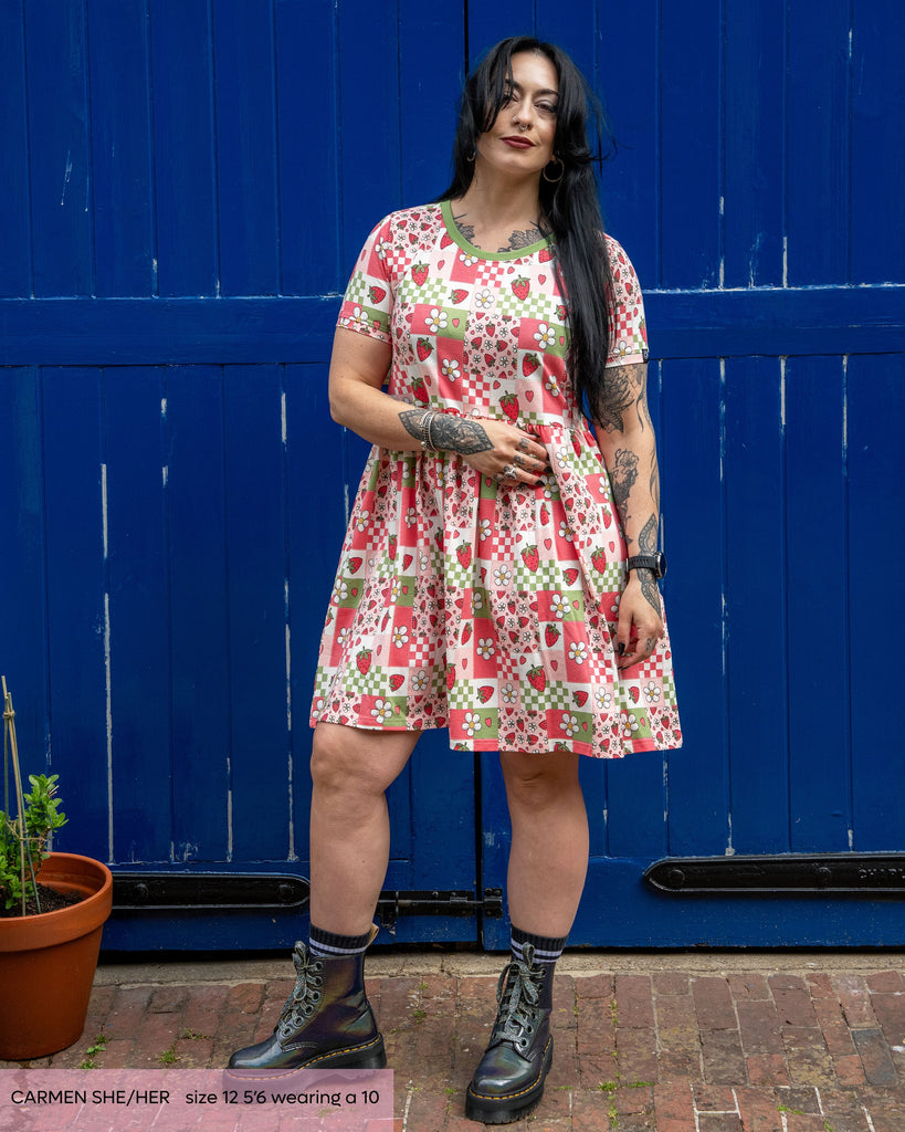 Tattooed gothic model, Carmen, wears the rebel romance strawberry picnic smock dress with grey platform boots outside in front of a blue wooden door. The oversized dress has a picnic style print in sage green, pastel pink, white and pastel red in colourblocks with strawberries, blossoms and checkerboard. 