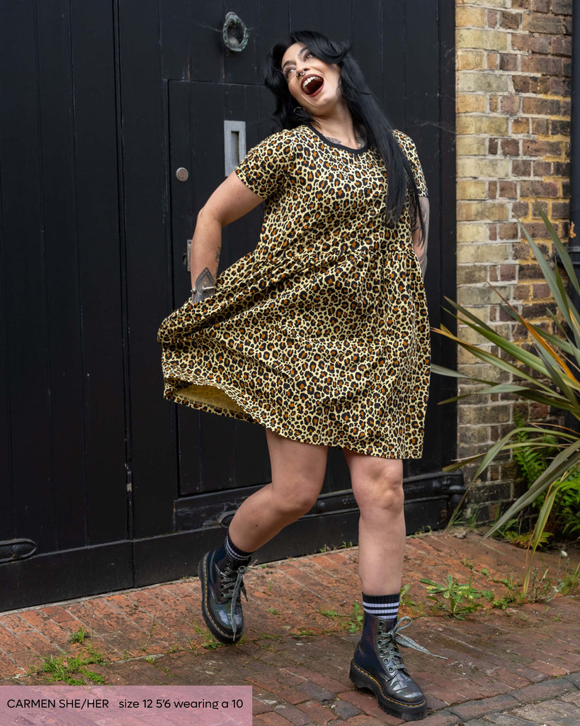 Carmen, a goth model with long black hair wearing the rebel romance leopard print smock dress with silver boots. She is taking a step forward swishing the dress out with her hands in the pockets and throwing her head back laughing. She is outside in a cobbled courtyard with a black door behind her. 