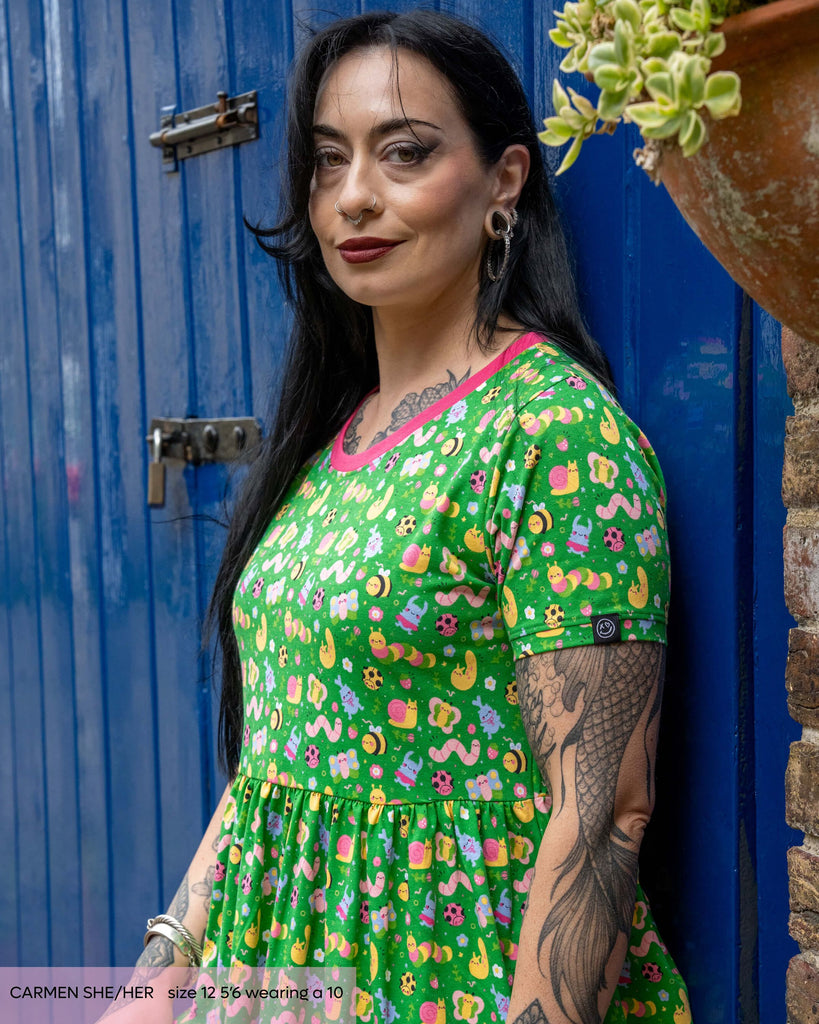 Alternative gothic tattooed woman, Carmen, wearing the rebel romance x dipstick bugs printed smock dress with contrasting pink neck line. The green base print has purple, yellow, pink and green bugs with cute faces all over.
