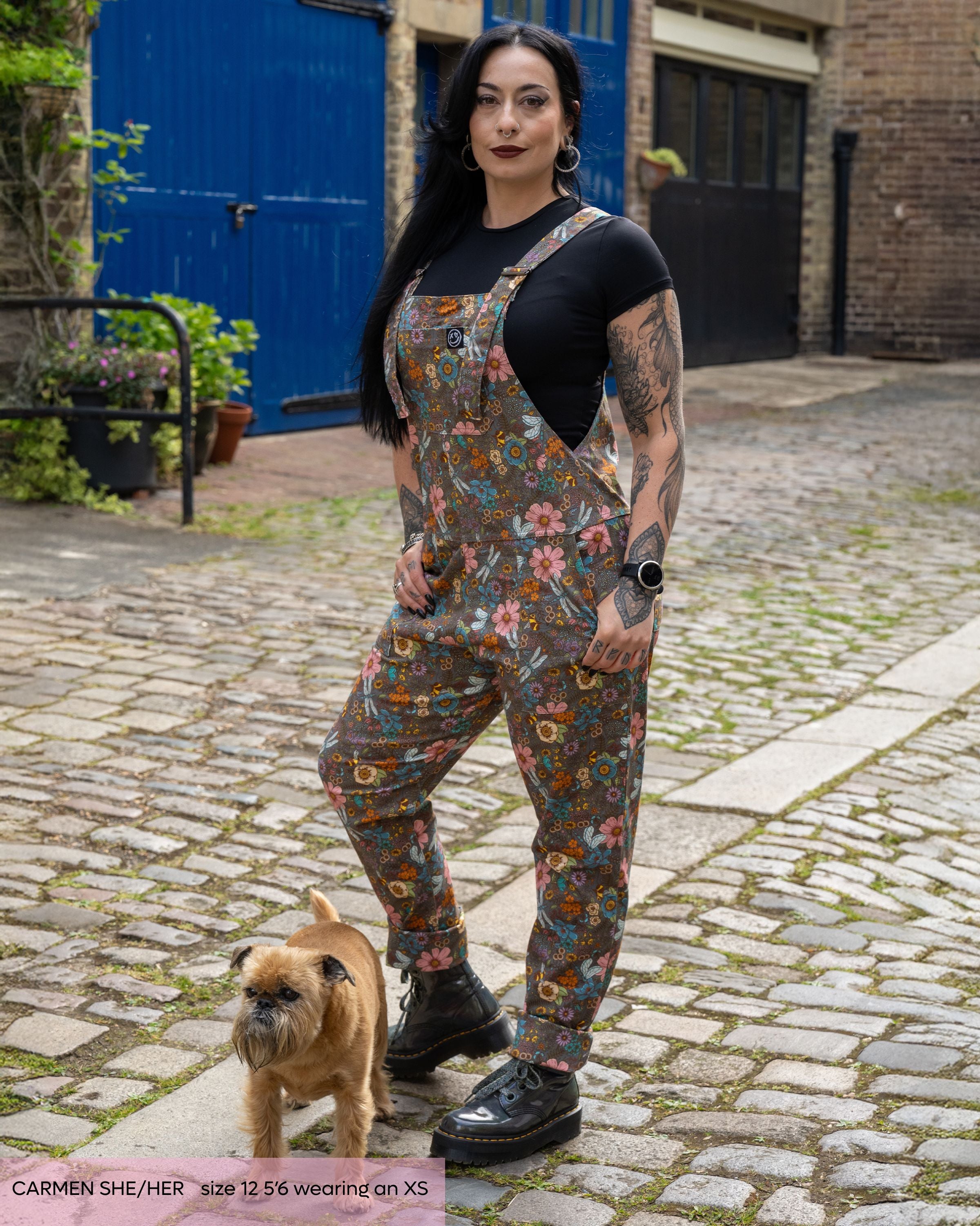 An alternative goth model, Carmen, is wearing the rebel romance cottagecore dungarees with a black crop top and grey platform boots. She is facing forward posing with her hands in the pockets with a small brown dog by her feet. The print is a warm grey base covered in florals, bees, dragonflies and butterflies.