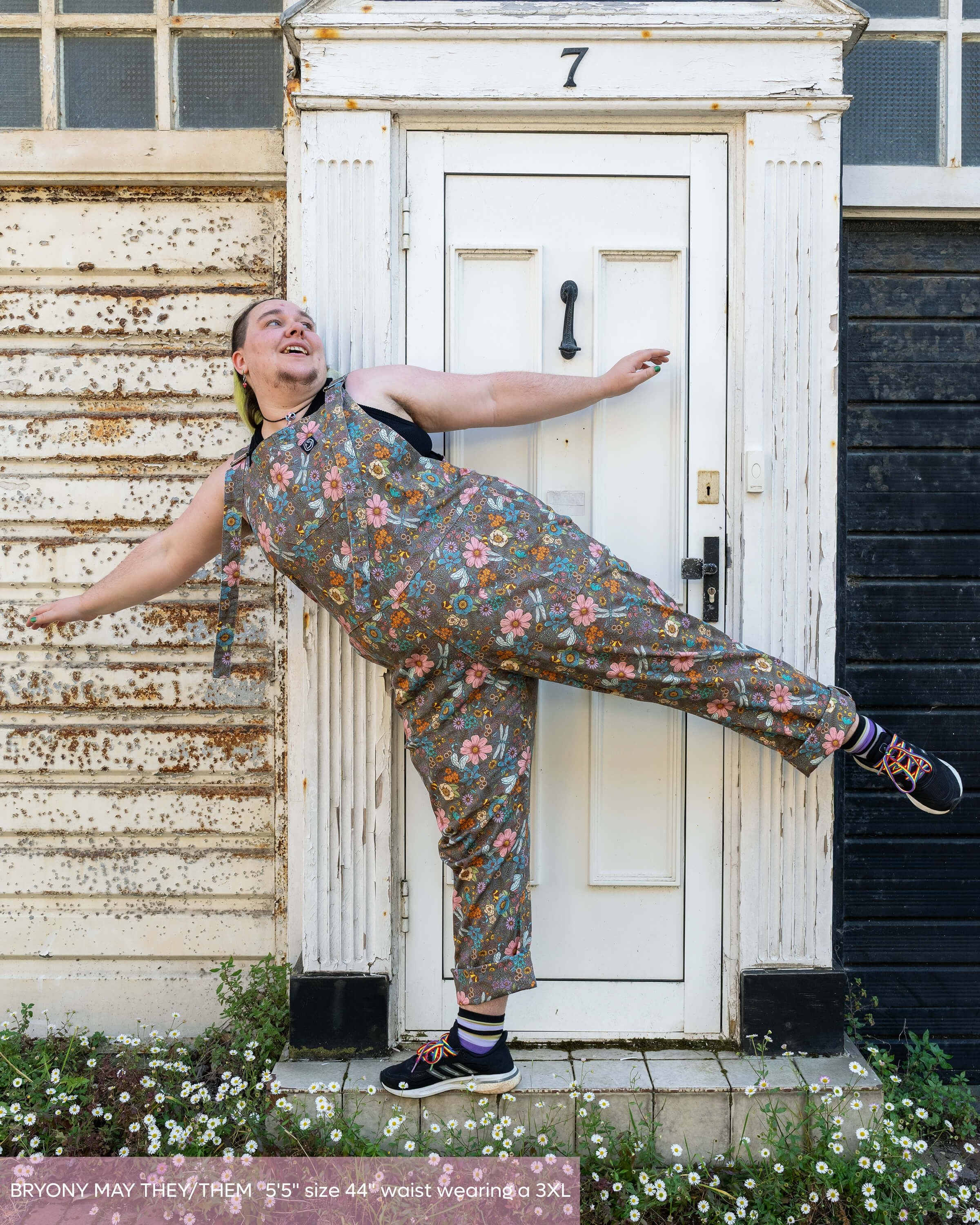 Bryony May, a model with long green dyed hair posing in a fun way balancing on a doorstep laughing wearing the cottagecore dungarees with rainbow laced  trainers and black crop top. The dungarees are a warm grey base colour with various colourful florals and bees, dragonflies, butterflies.