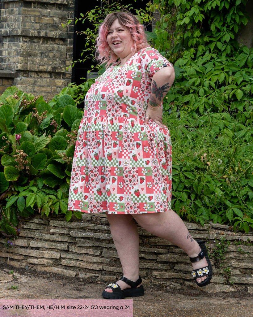 Non binary model, Sam, wearing the strawberry picnic smock dress with daisy floral sandals. The dress features strawberries, blossoms and colour blocks in sage green, pink and pastel red. They are stood laughing kicking a leg out with their hands on their hips outside in front of a lush green bush.