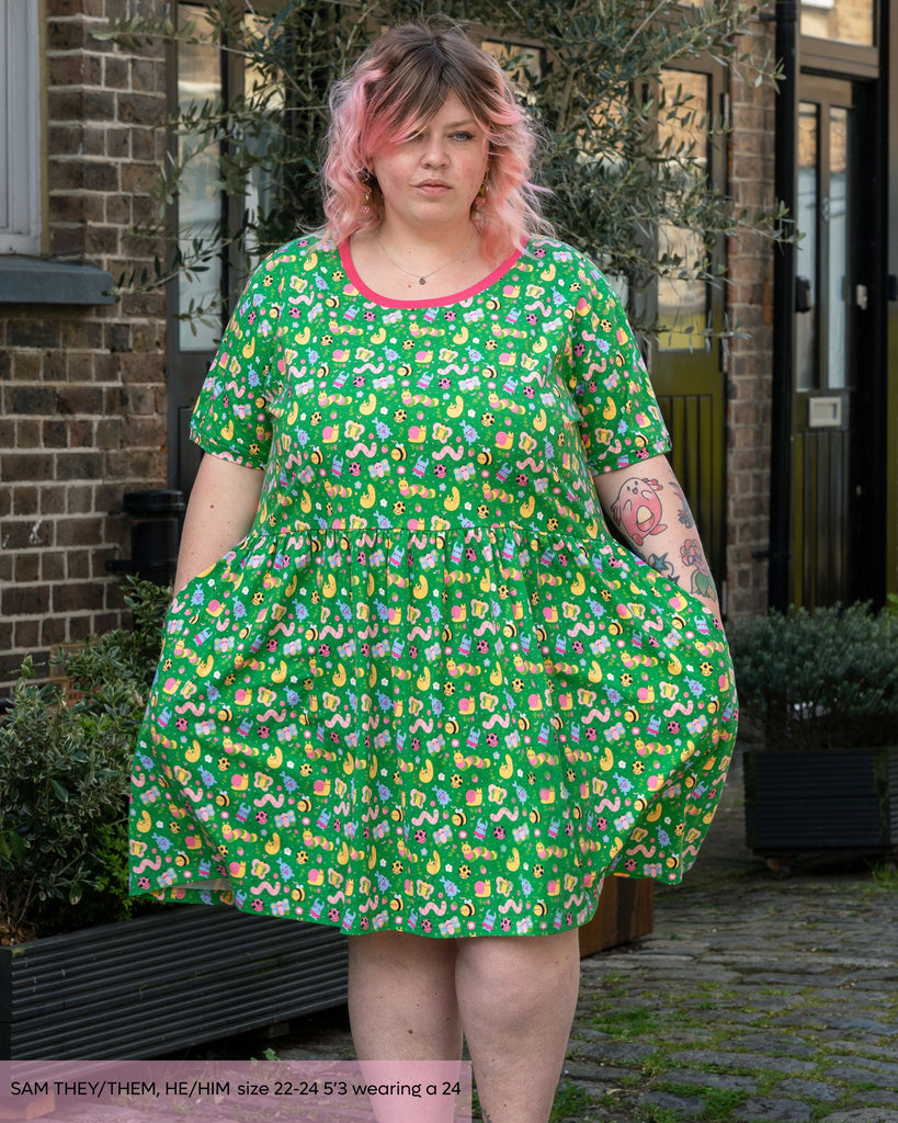 Sam, a non binary tattooed model with pink curly hair wearing the rebel romance x dipstick bugs smock dress. The dress print is a bright spring green base with kawaii bugs such as caterpillars, slugs, stag beetles, ladybirds, butterflies and more in pinks, yellows, blues and purple. 