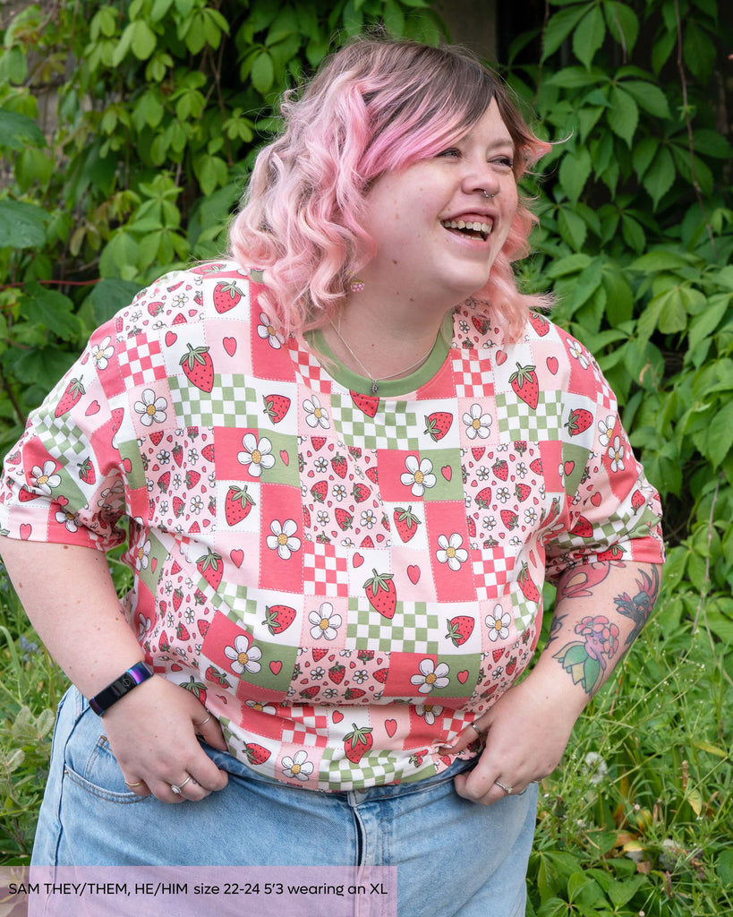 Sam, a non binary masc model with pink curly hair wearing the strawberry picnic crop top with light blue denim trousers. They are laughing leaning forward looking right. The print features sage green, pastel pink, strawberries, hearts and white blossoms. 