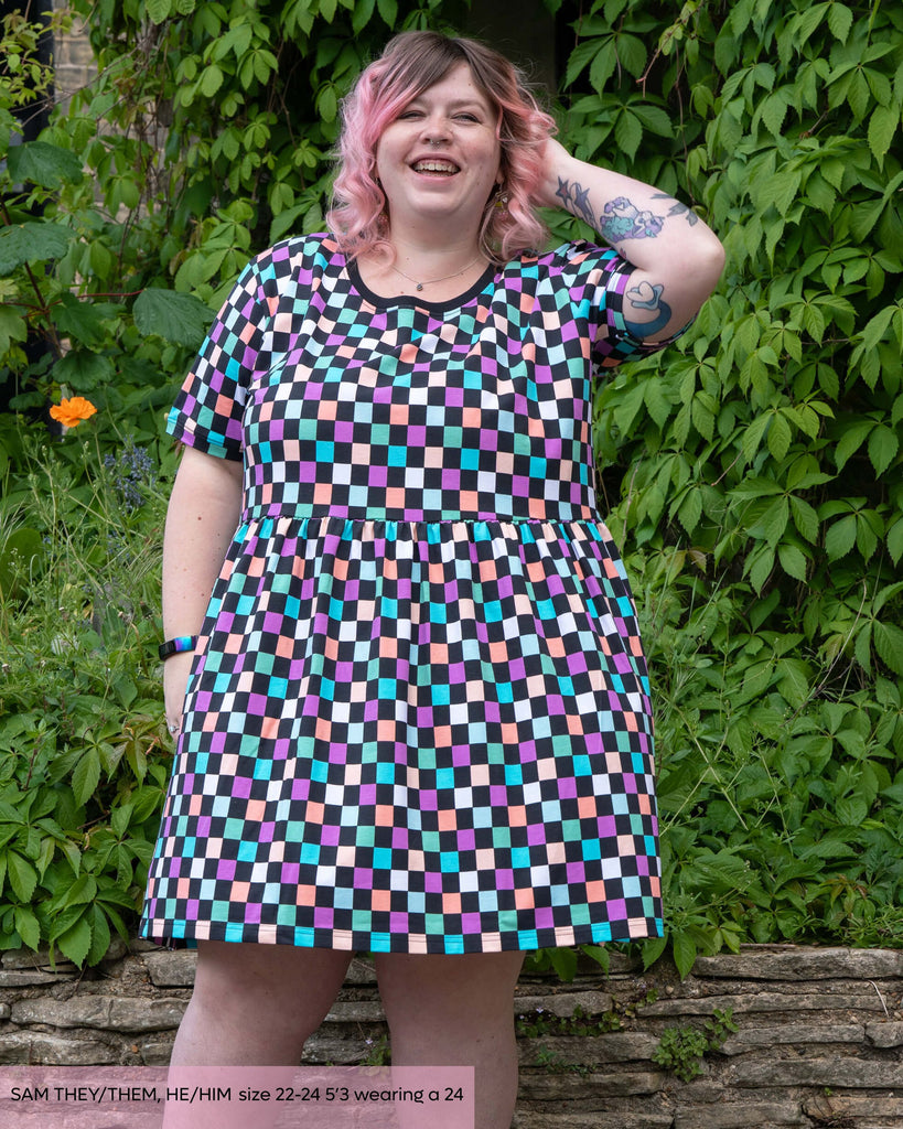 Sam, a non binary model with short pink curly hair and tattoos laughing stood outside in front of a lush green foliage wall wearing the rebel romance checkerboard smock dress. The dress is a black and colourful checkered print with a contrasting black neckline. 
