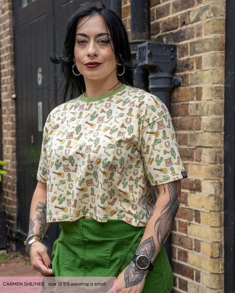 Carmen, a gothic tattooed woman wearing the cowboy oversized boxy crop tee with the Rebel Romance green dungarees. The butter yellow pastel print features cowboy themed illustrations by Yasmin Cousins. She is smiling facing forward with a brick wall behind them. 