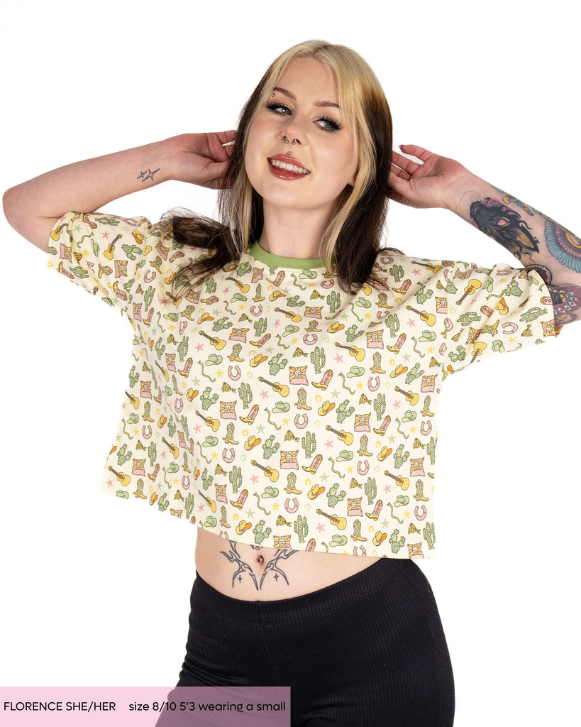 Florence, a tattooed alternative woman wearing the rebel romance oversized cowboy boxy crop tee designed by Yasmin cousins. The butter yellow print with a sage green contrasting trim features cowboy themed illustrations of cacti, guitars, cowboy hats, horseshoes, snakes and more. She's paired it with black leggings and is smiling in front of a white background. 